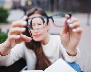 Closeup portrait of young women with glasses. She has eyesight problems and is squinting his eyes a little bit. Beautiful girl is holding eyeglasses right in front of camera with two hand