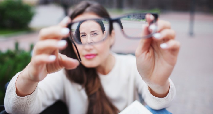 Closeup portrait of young women with glasses. She has eyesight problems and is squinting his eyes a little bit. Beautiful girl is holding eyeglasses right in front of camera with two hand